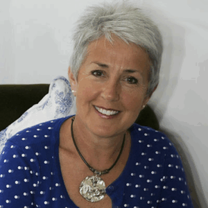 Head and shoulder photograph of a smiling lady. She has short grey hair and is sat down wearing a blue cardigan with white spots and a shell necklace.